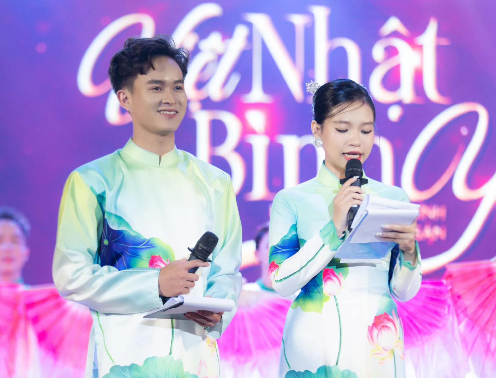 Two MCs in lotus-print áo dài hosting in front of the Việt Nhật Bình Y stage backdrop