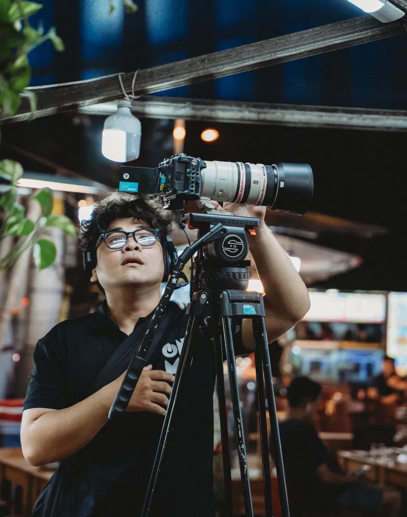 Cinematographer lining up a telephoto shot on a tripod at a night food market