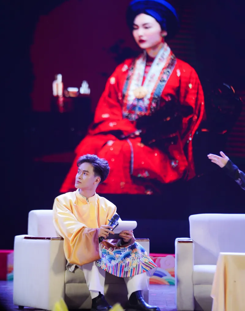 Speaker in a golden áo tấc seated on stage, a Nhật Bình portrait glowing on the screen behind