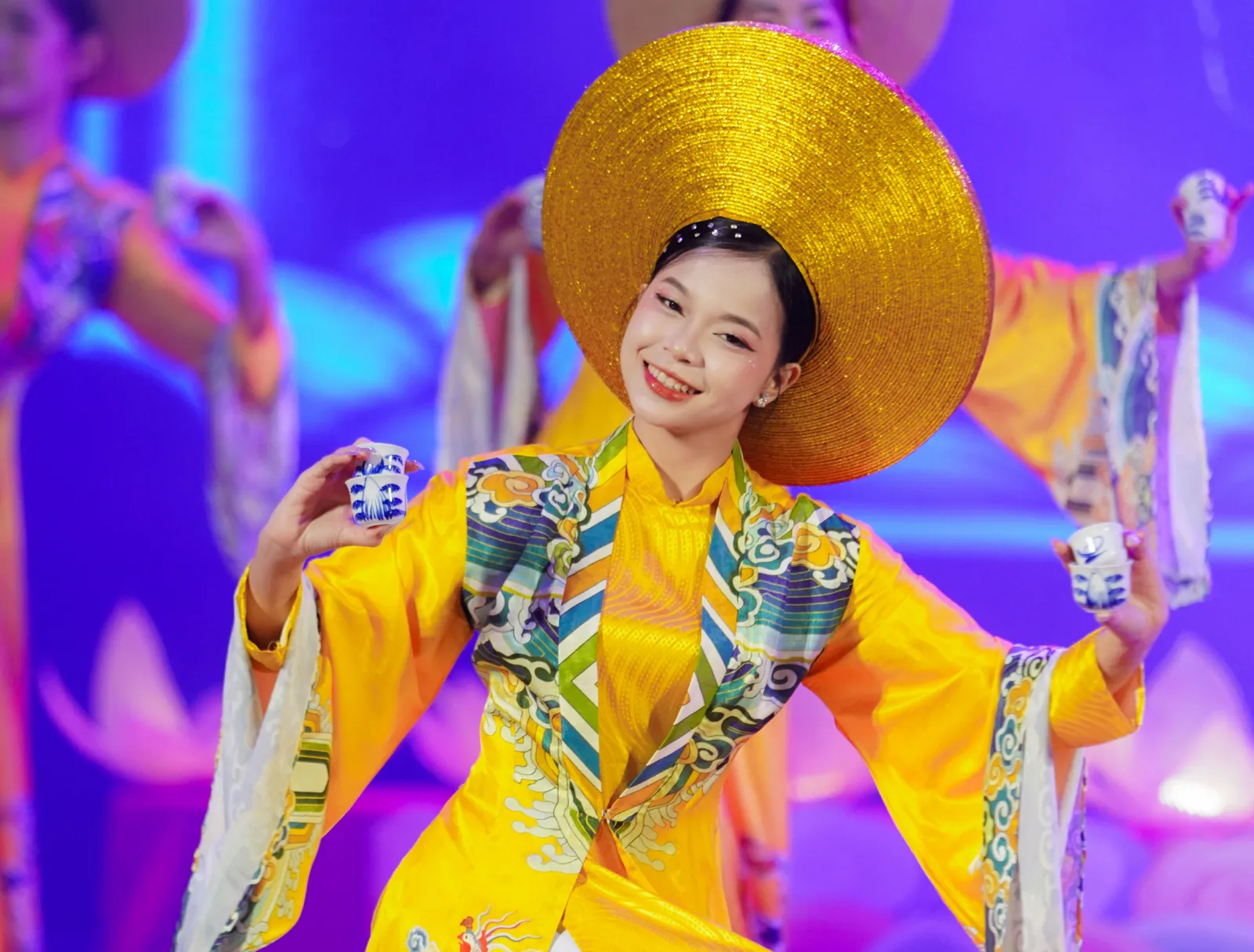 Dancer in a royal-yellow costume and wide golden hat balancing porcelain teacups