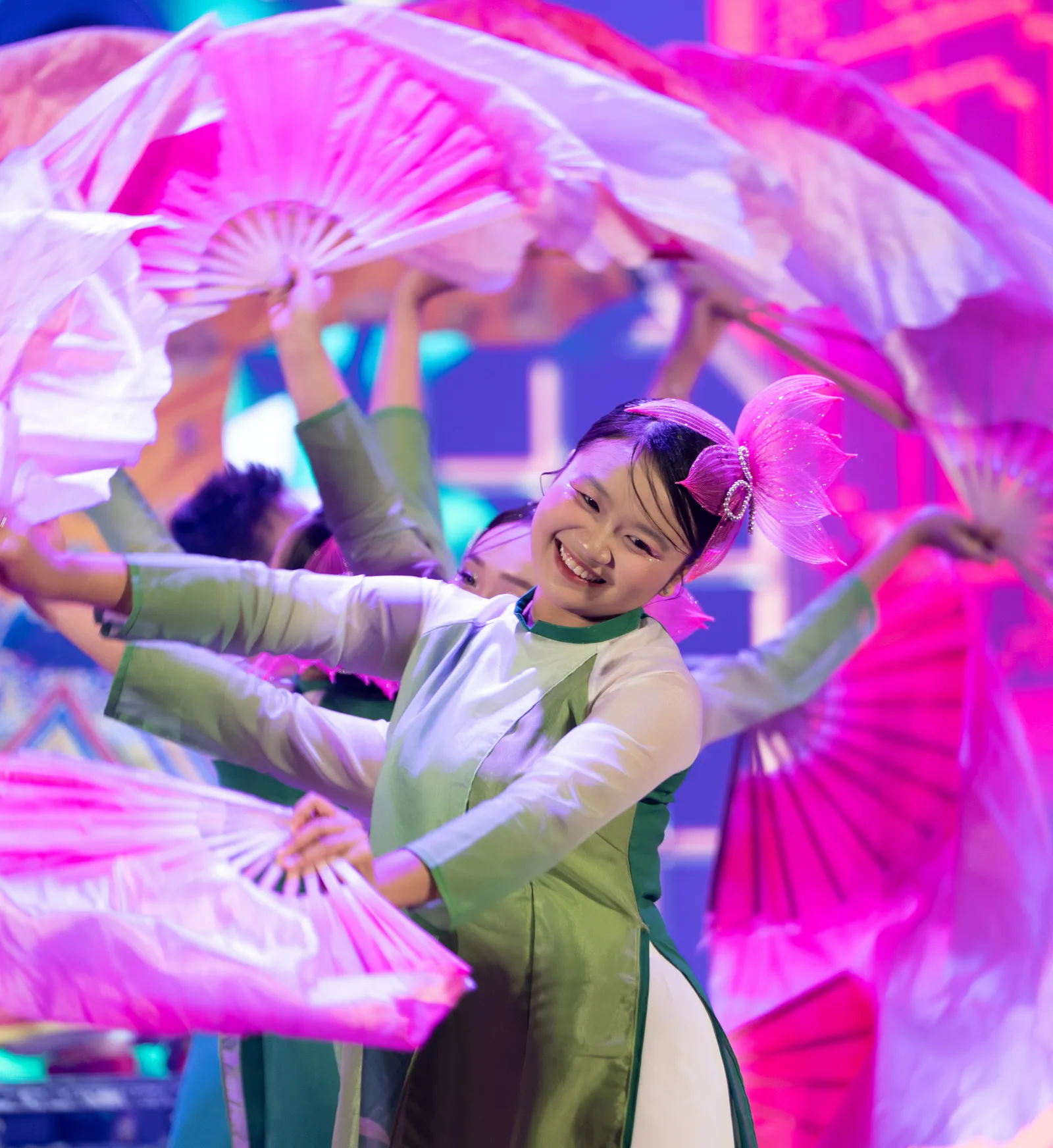 Dancer in a green áo dài smiling mid–fan dance, surrounded by swirling pink fans